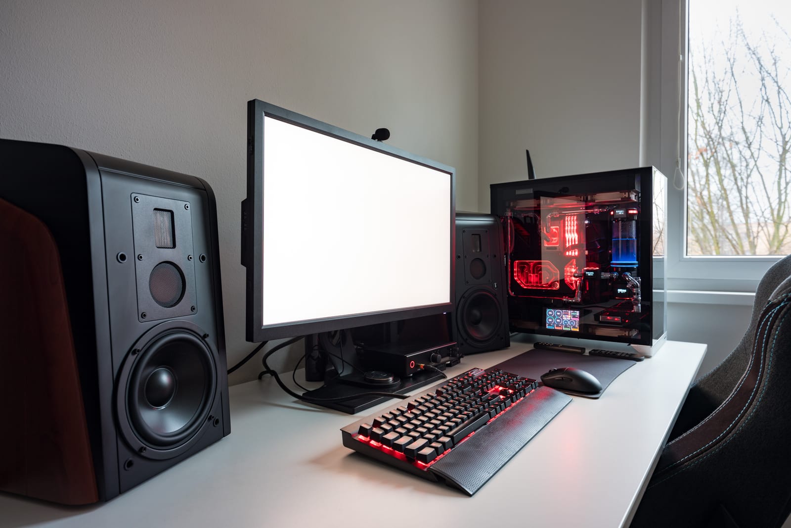 A modern gaming setup on a white desk, with the gaming monitor as the centerpiece, flanked by a high-end PC with RGB lighting and large speakers.